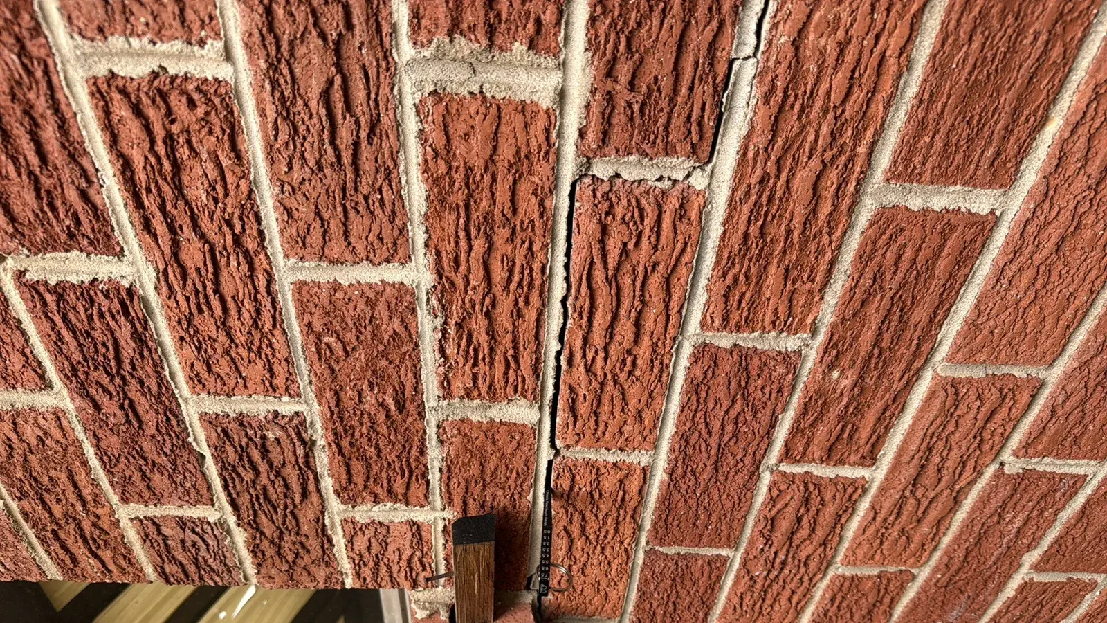 Vertical crack stepping through the mortar joints of a red textured brick wall, with a crack gauge at the base of the crack.