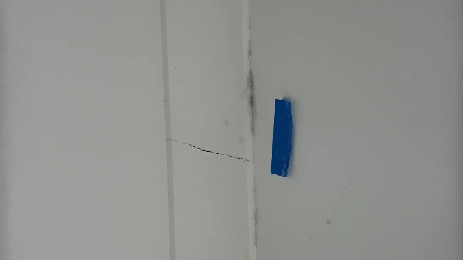 Dark mould spotting along a joint in a white painted ceiling lining, beside a hairline crack and a strip of blue marking tape